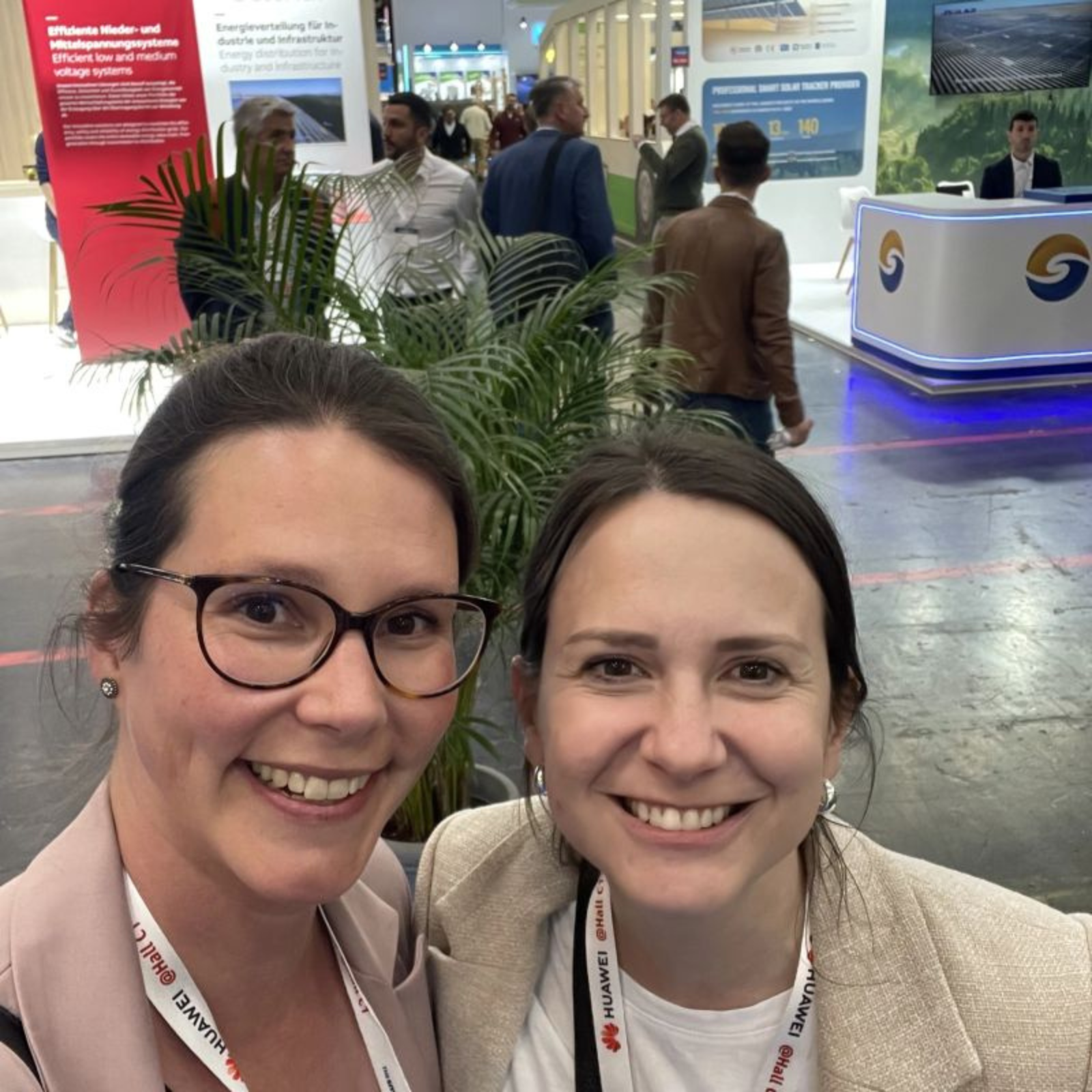 Two smiling women with conference lanyards take a selfie at a trade show with people and booths behind.