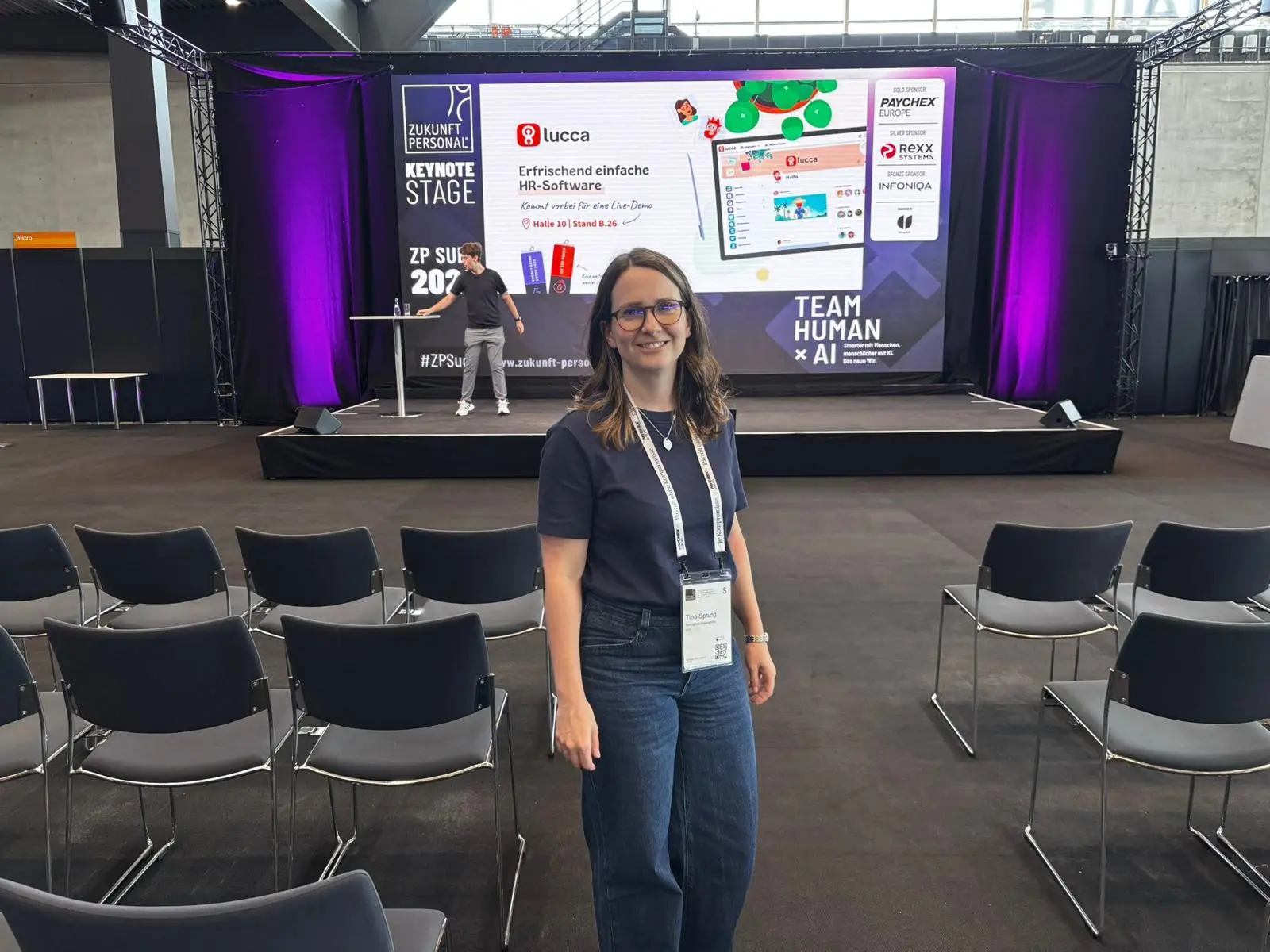 Woman with glasses and badge standing smiling in front of empty chairs at a conference stage with purple lights.