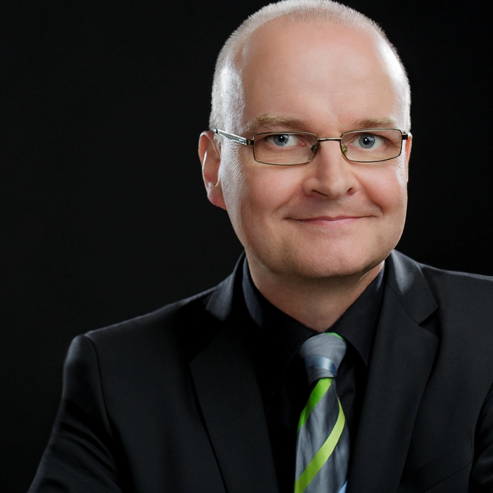 Bald man in glasses wearing black suit and striped tie smiling against a black background.
