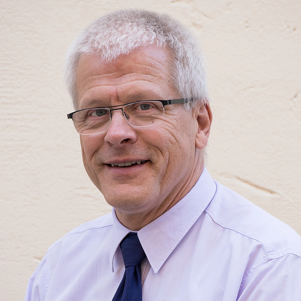 Smiling older man with gray hair and glasses wearing a light purple shirt and dark blue tie.