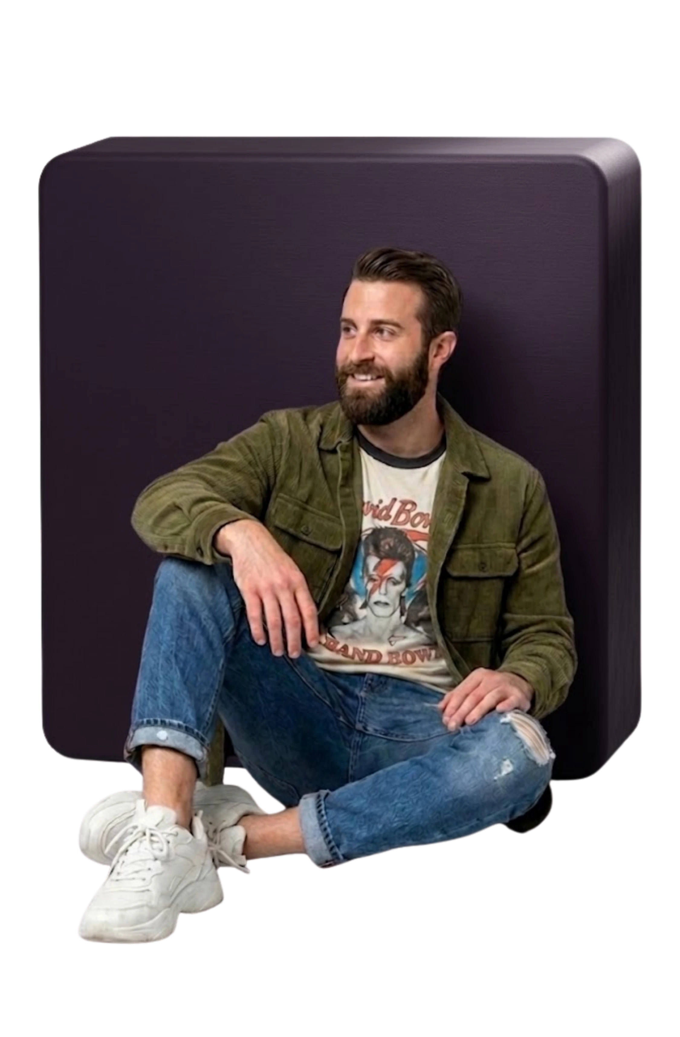 Bearded man sitting cross-legged wearing a green jacket, David Bowie shirt, jeans, and white sneakers.