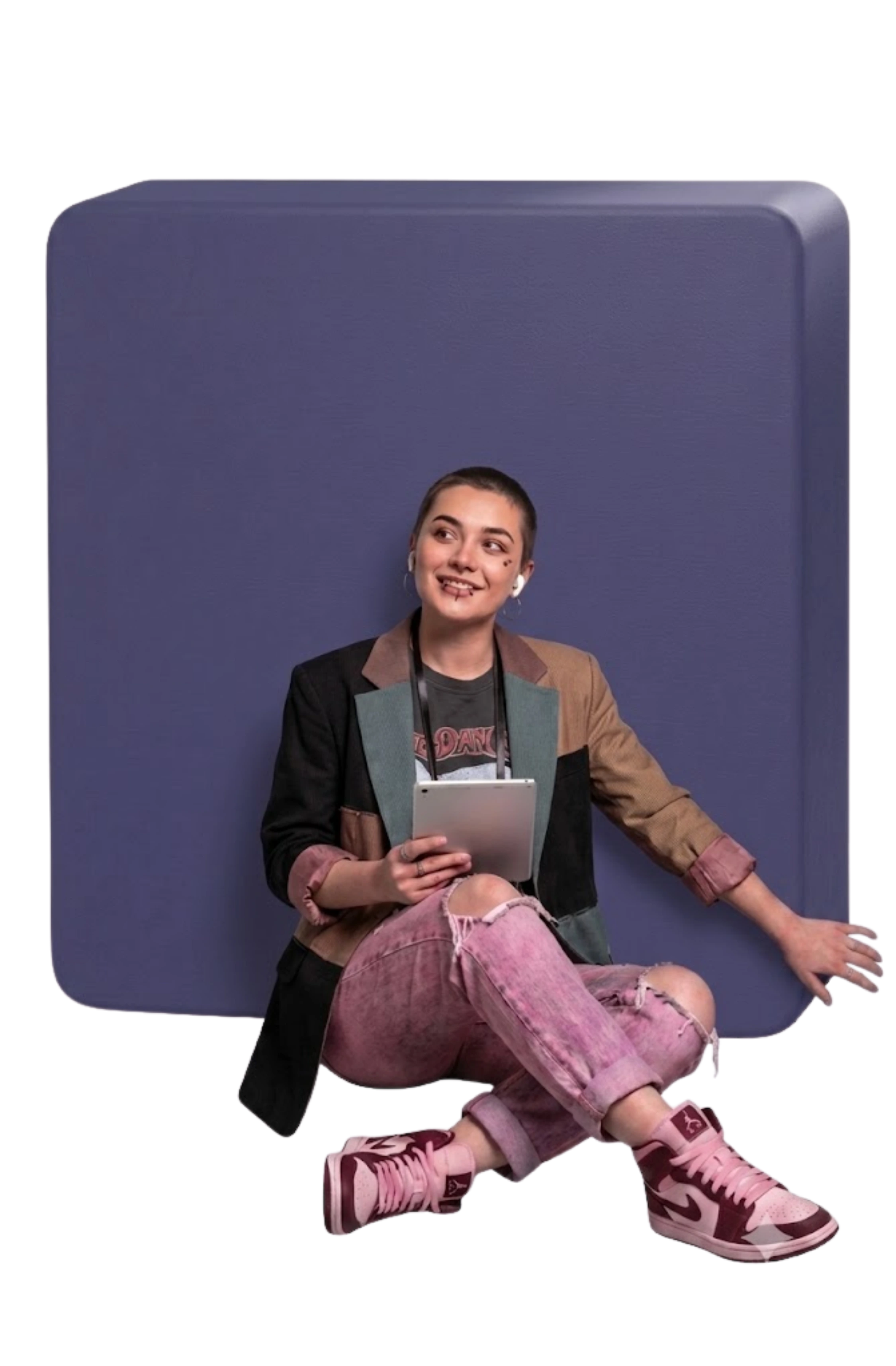 Smiling young person with short hair, pierced lips, AirPods, colorful blazer, and pink ripped jeans holding a tablet.