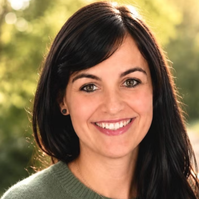 Smiling woman with long dark hair wearing green sweater outdoors with blurred green background.