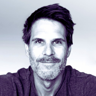 Portrait of a man with short dark hair and a beard, smiling slightly against a light background.