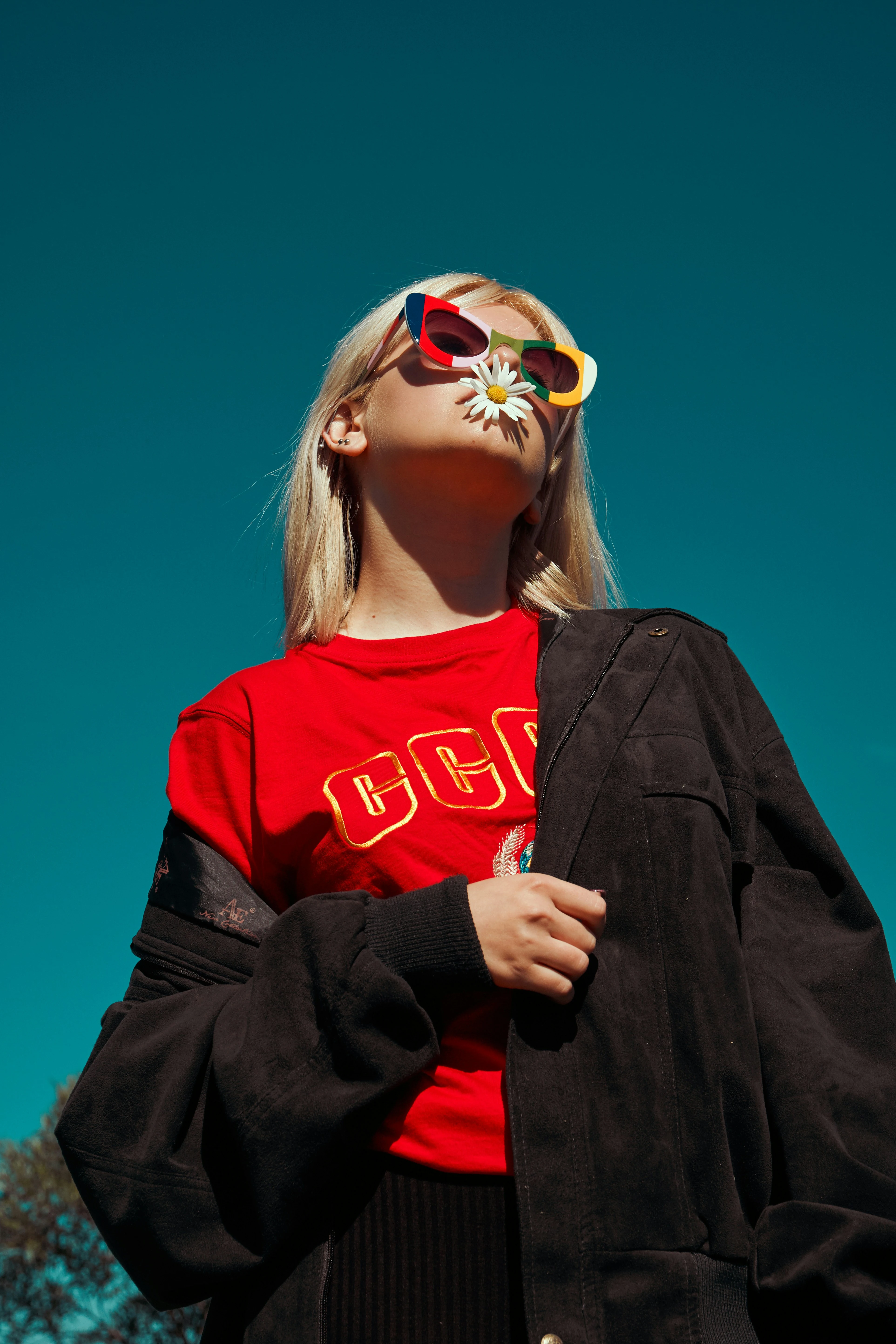 Woman wearing colorful sunglasses with a daisy held in her mouth against a clear blue sky.