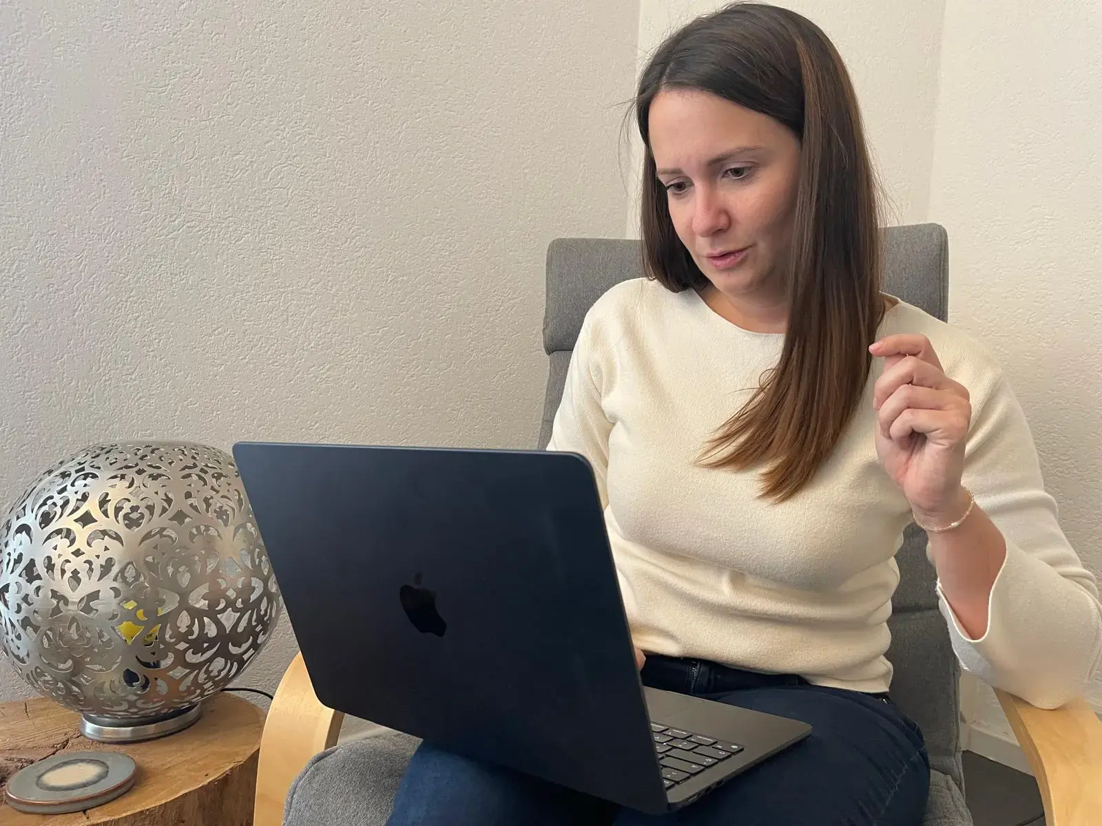Woman in white sweater sitting on chair, focused on a black laptop on her lap.