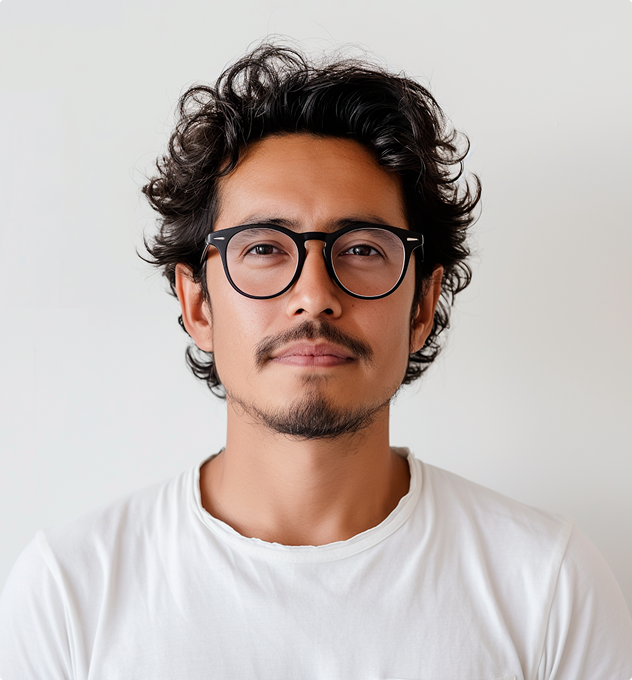 Portrait of a man with curly black hair, a mustache and goatee, black round glasses, wearing a white t-shirt, against a plain light background.