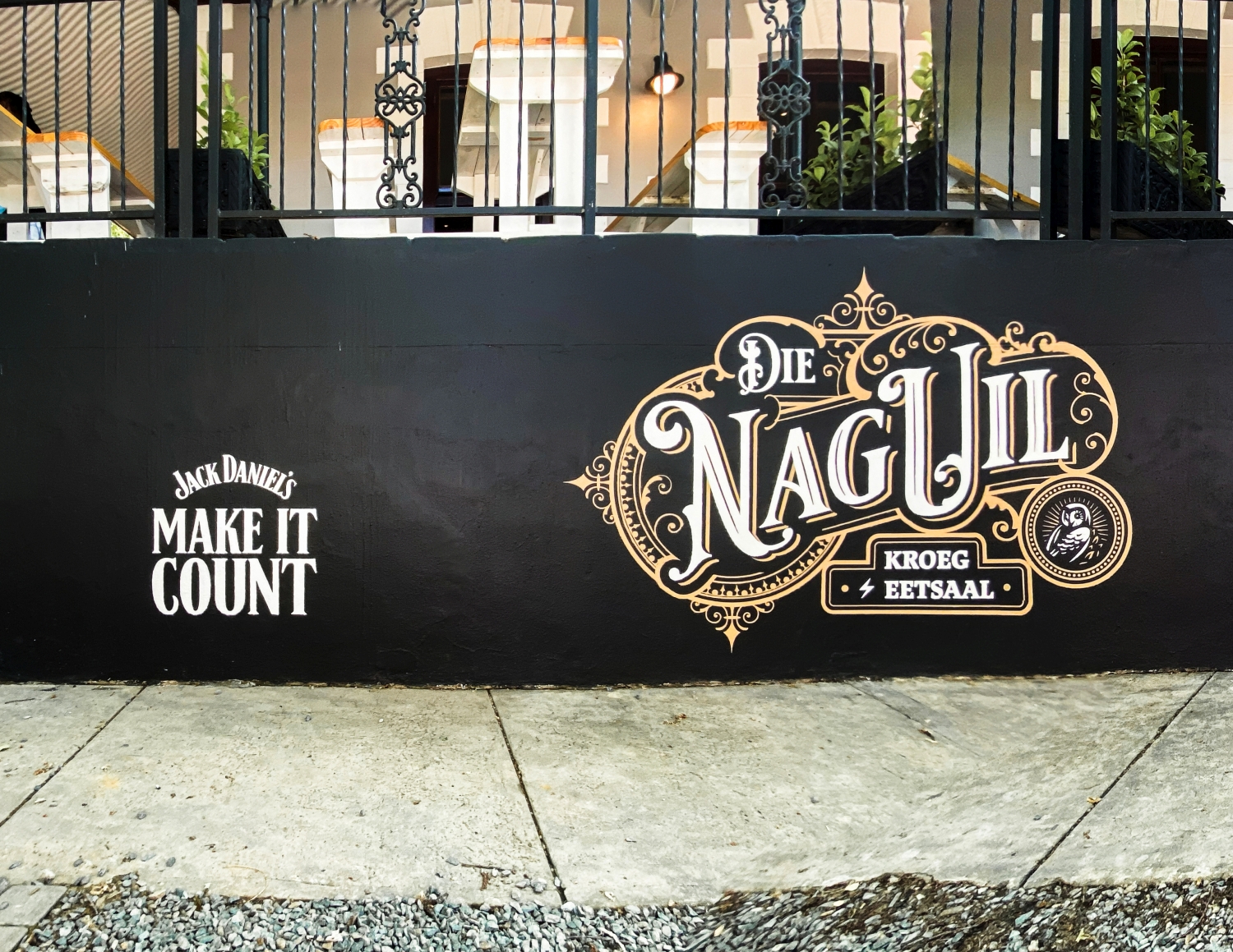Black wall with white and gold signage displaying 'Jack Daniel's Make It Count' and 'Die Nagu III Kroeg Eetsaal' under a balcony with metal railings.