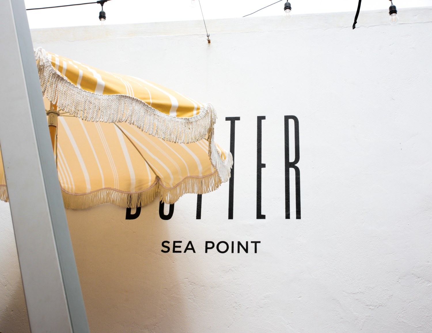 Yellow and white striped outdoor umbrella with fringe next to a white wall with the text 'BUTTER SEA POINT'.