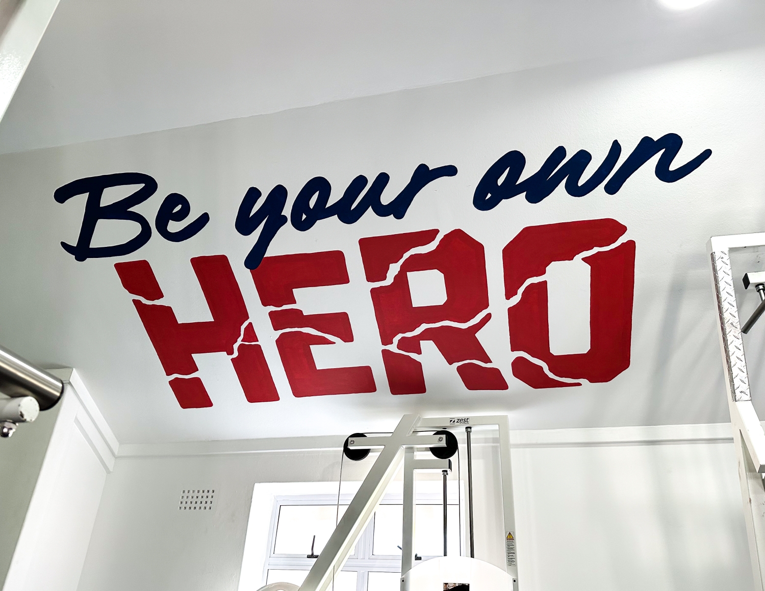 Motivational phrase "Be your own HERO" painted on a white gym wall above exercise equipment.