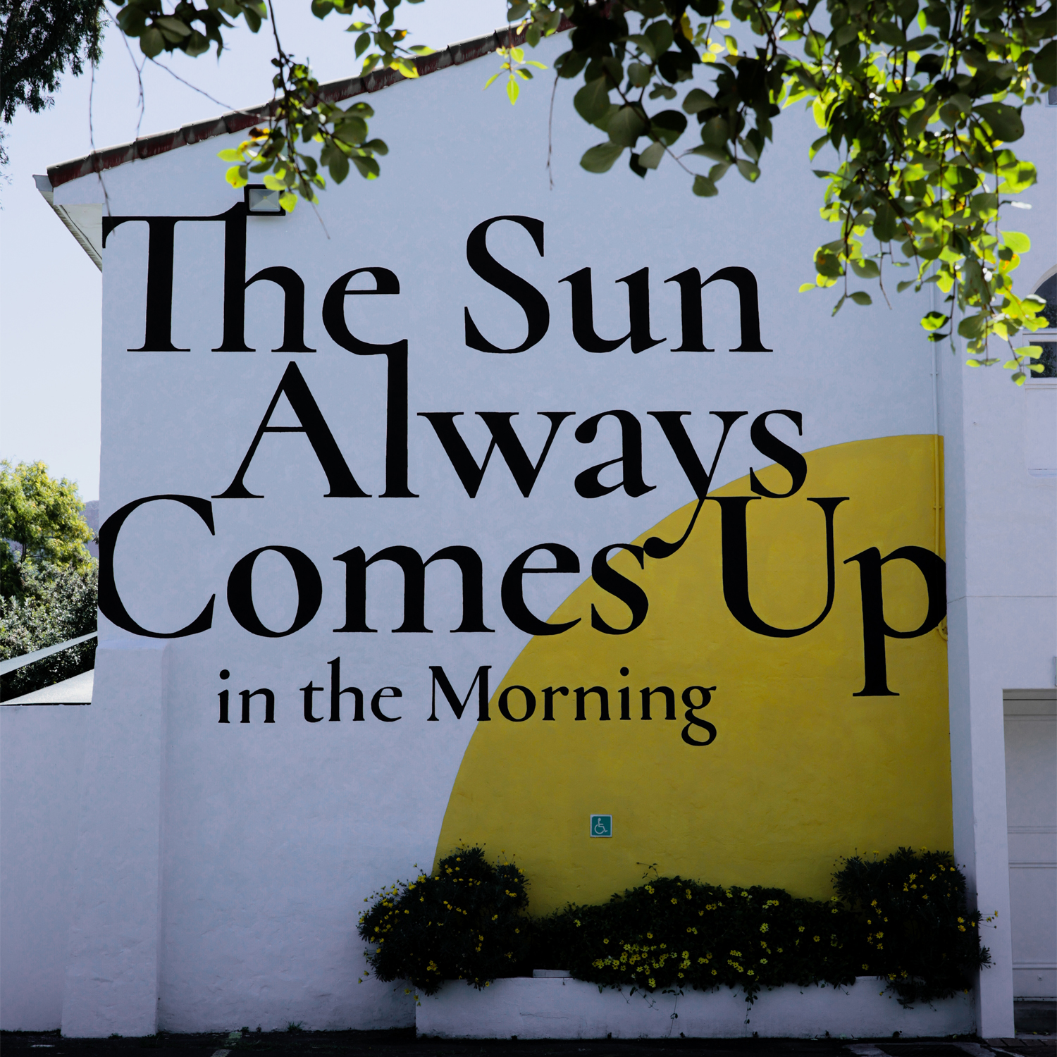 White wall mural with large black text reading 'The Sun Always Comes Up in the Morning' over a yellow semicircle, surrounded by plants and tree branches.