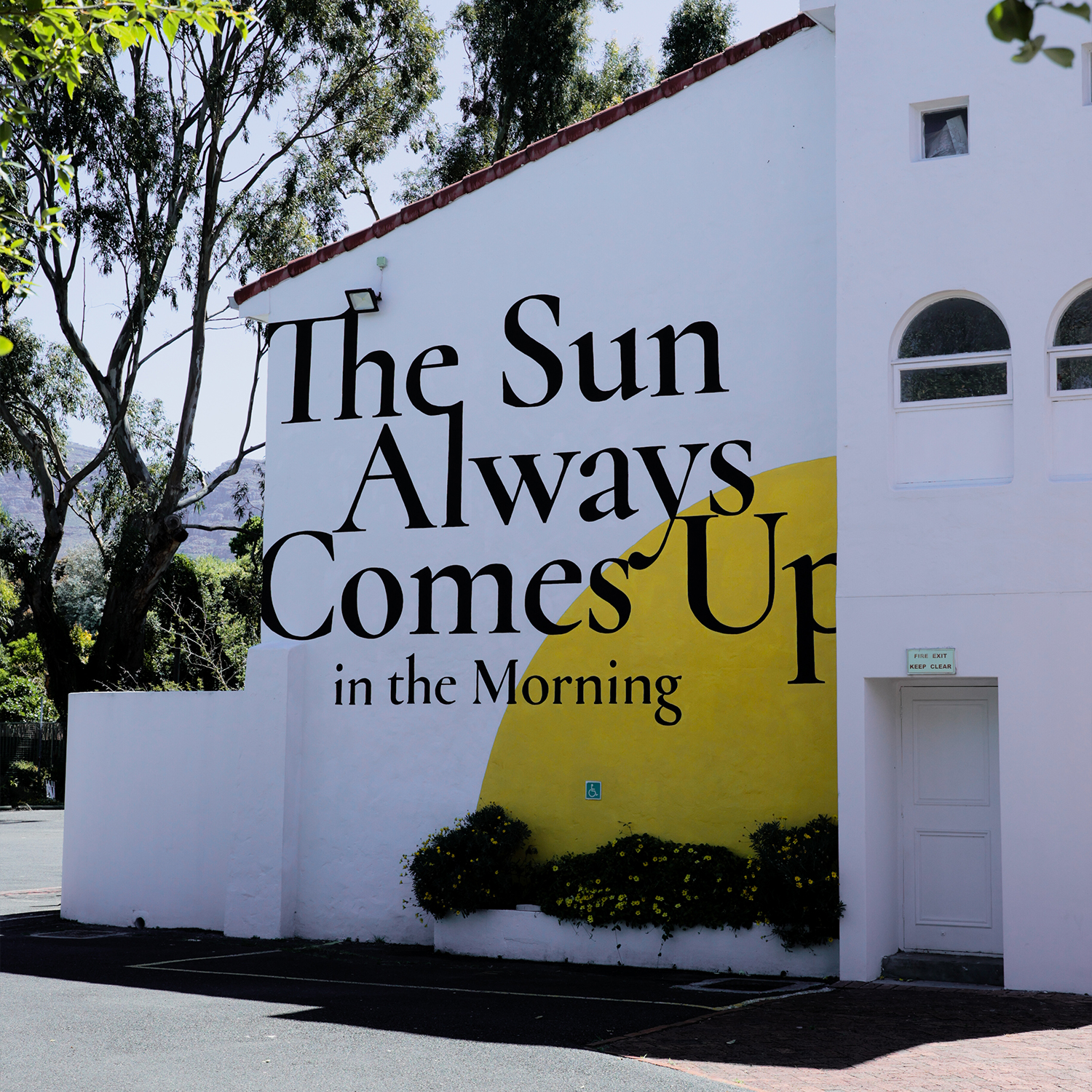 White building wall with a large mural saying 'The Sun Always Comes Up in the Morning' and a half yellow sun graphic above green bushes.