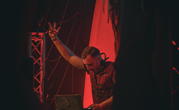 DJ performing with raised hand and focused expression in red-lit venue with laptop in front.