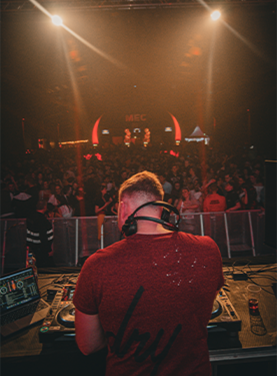 DJ wearing headphones performing on stage with a crowd and bright stage lights in the background.