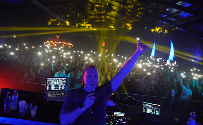 DJ holding a microphone and phone with flashlight on, performing in front of a large crowd at a nightclub with colorful lighting.