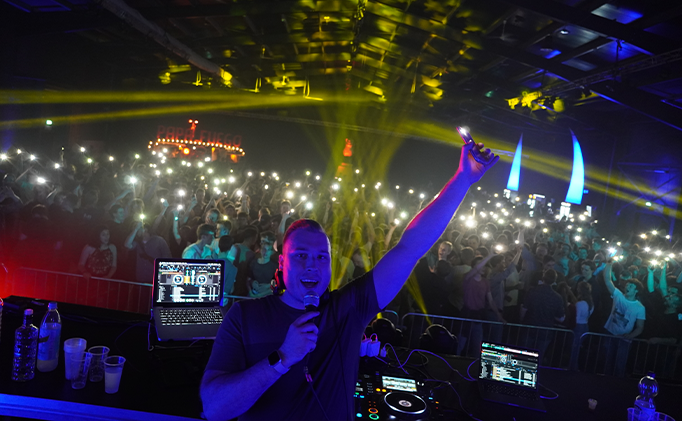 DJ holding a microphone and phone with flashlight on, performing in front of a large crowd at a nightclub with colorful lighting.