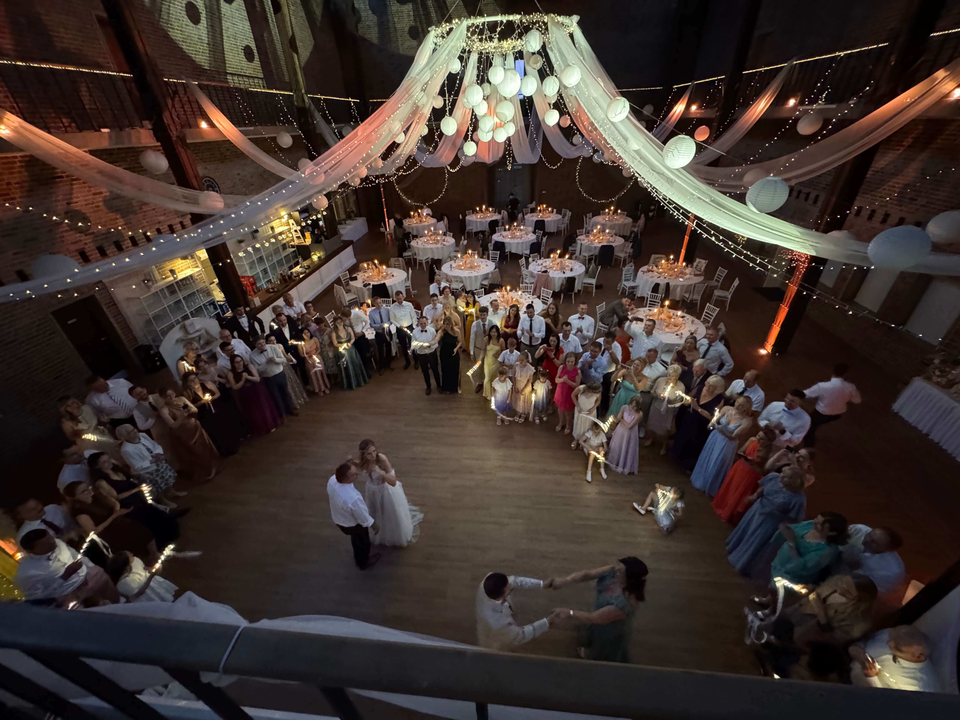 Overhead view of a wedding reception with guests standing in a circle around a bride and groom on the dance floor, with string lights and hanging decorations above.