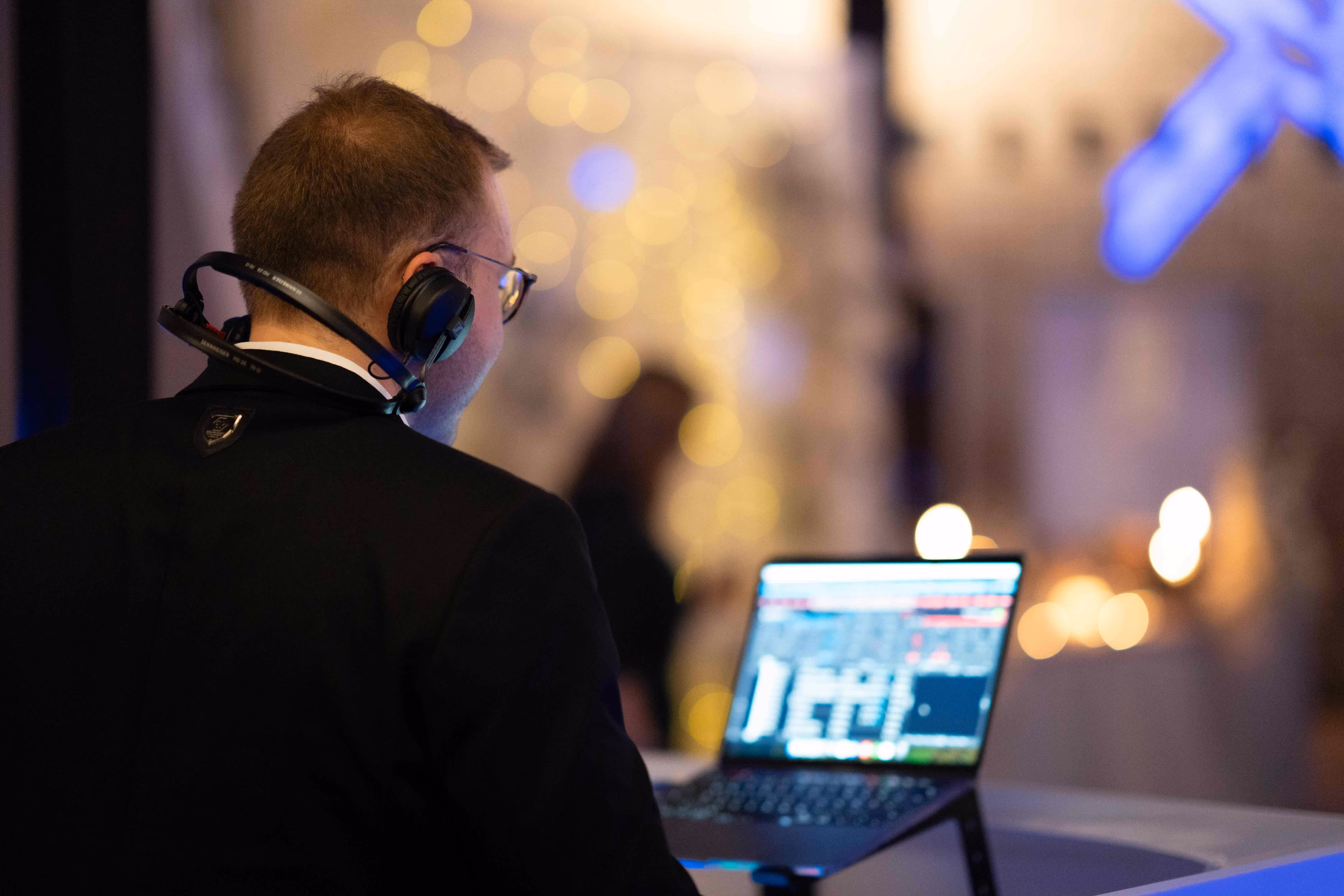 Man wearing headphones and glasses working on a laptop with a blurred background of lights.