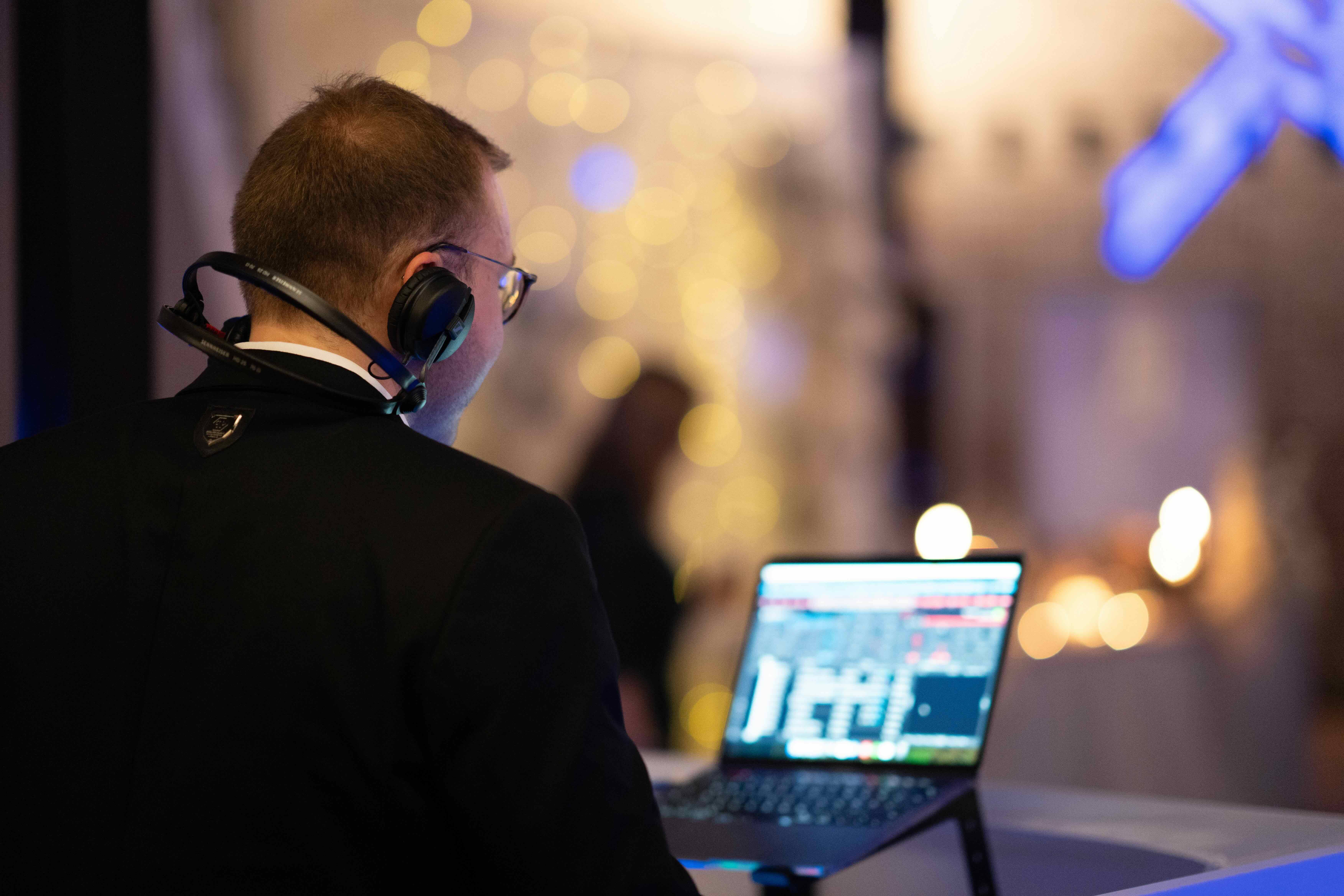 Man wearing headphones and glasses working on a laptop with a blurred background of lights.
