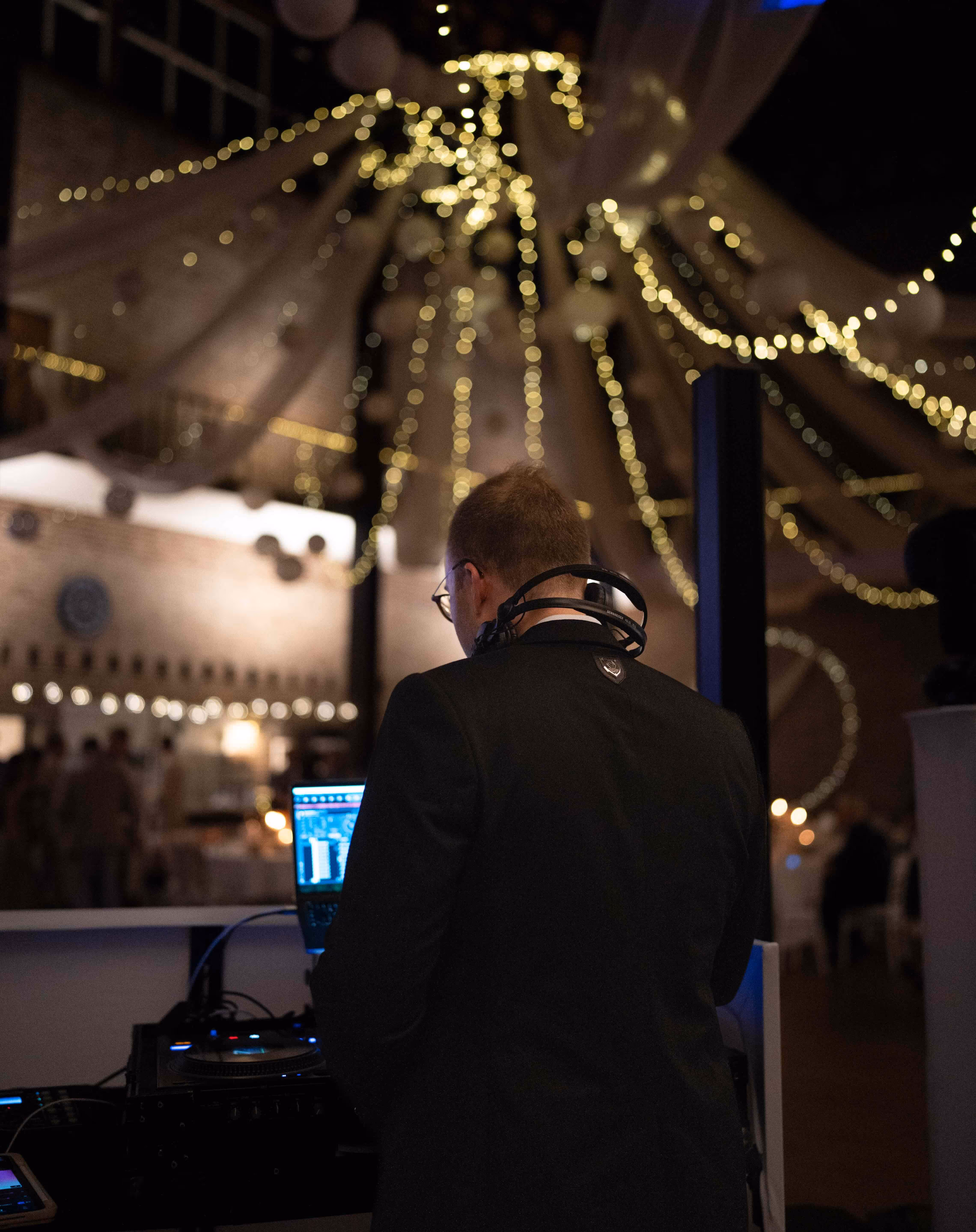 DJ in a black suit working on a laptop with headphones around his neck under warm string lights at an indoor event.