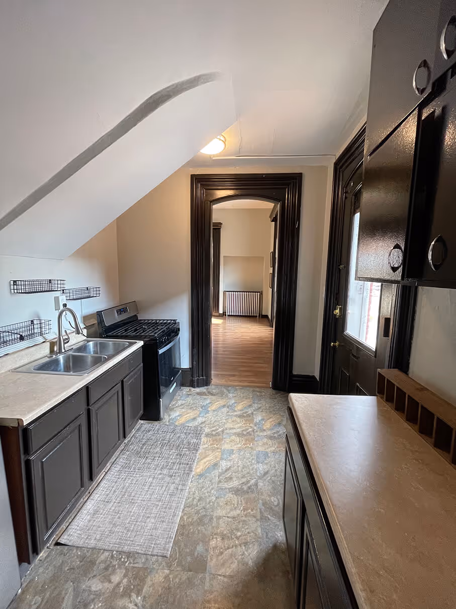 compact galley kitchen under a slanted ceiling featuring dark wood cabinets, a stainless steel sink and black stove, tiled floor, and a doorway leading to a room with wood flooring