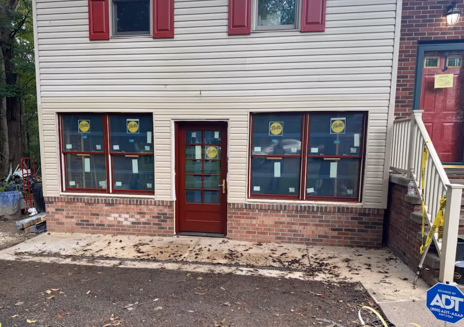 front of a house with beige siding and brick lower walls, featuring newly installed red‑framed windows and a red door, with caution tape on the neighboring stair railing and an ADT security sign