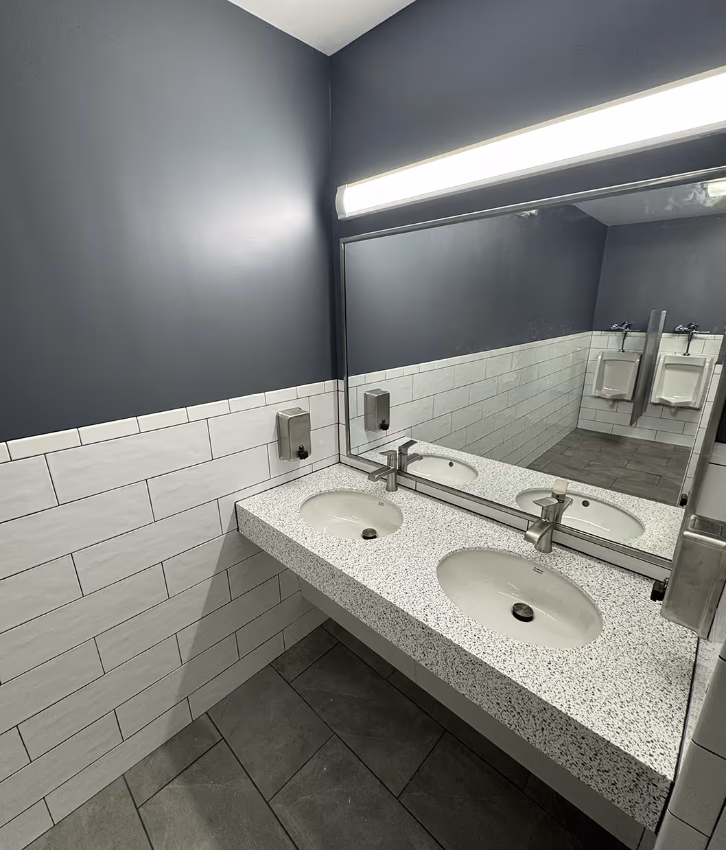 modern commercial bathroom vanity featuring a speckled two‑basin countertop with stainless faucets and dispensers, a large mirror reflecting urinals, white subway tile walls, and grey floor tiles