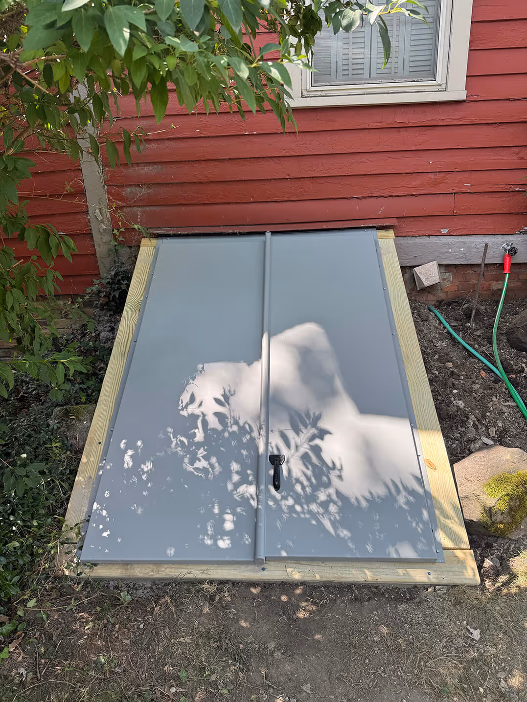 grey metal cellar doors set flush with the ground outside a red wooden house, framed with wood and surrounded by greenery and a garden hose