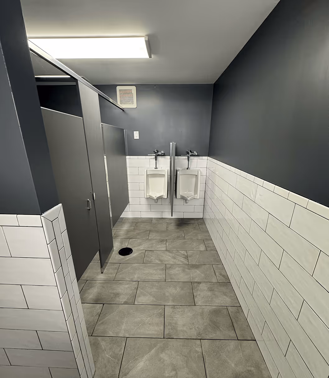 modern commercial restroom featuring dark walls, white subway tile wainscoting, grey floor tiles, and two wall‑mounted urinals separated by a privacy divider
