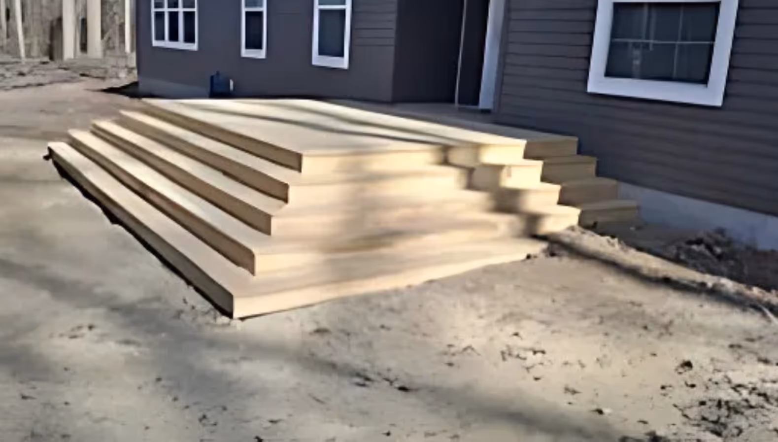 freshly built wide wooden deck steps leading up to a grey‑sided house with white‑trimmed windows