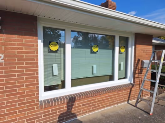 large triple‑pane window with a white frame newly installed in a brick wall, with yellow labels on the glass and a ladder leaning nearby