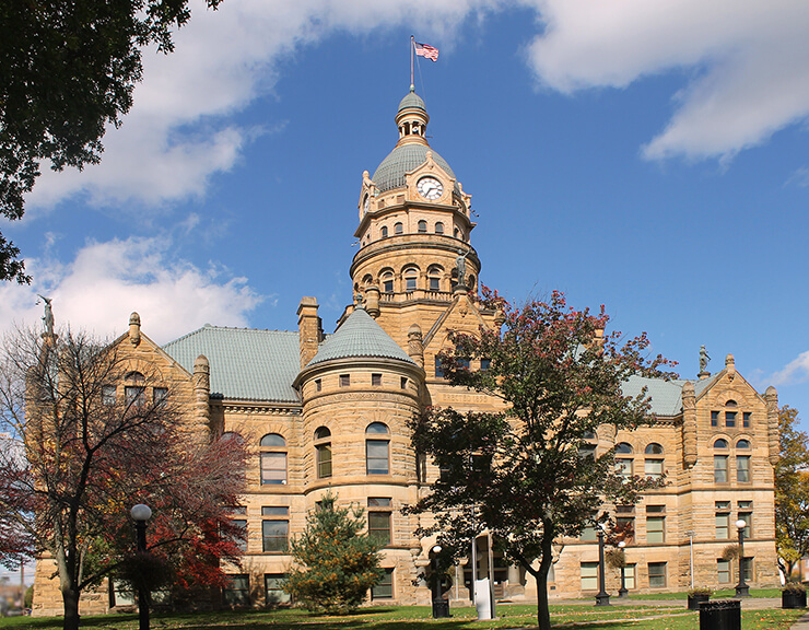 grand sandstone building with a green domed clock tower topped by an American flag, featuring arched windows and turrets, surrounded by trees under a partly cloudy sky