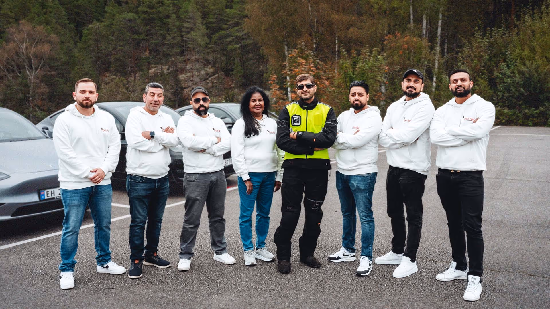 Åtte personer står på parkeringsplassen foran trær, de fleste i hvit hettegenser, én i svart og gul jakke.