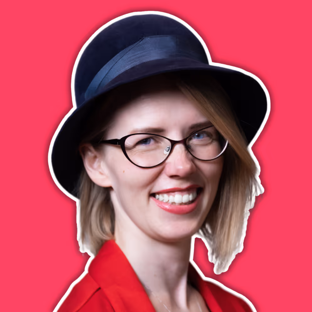 Smiling woman with light hair wearing glasses, a navy hat, and a red blazer against a pink background.