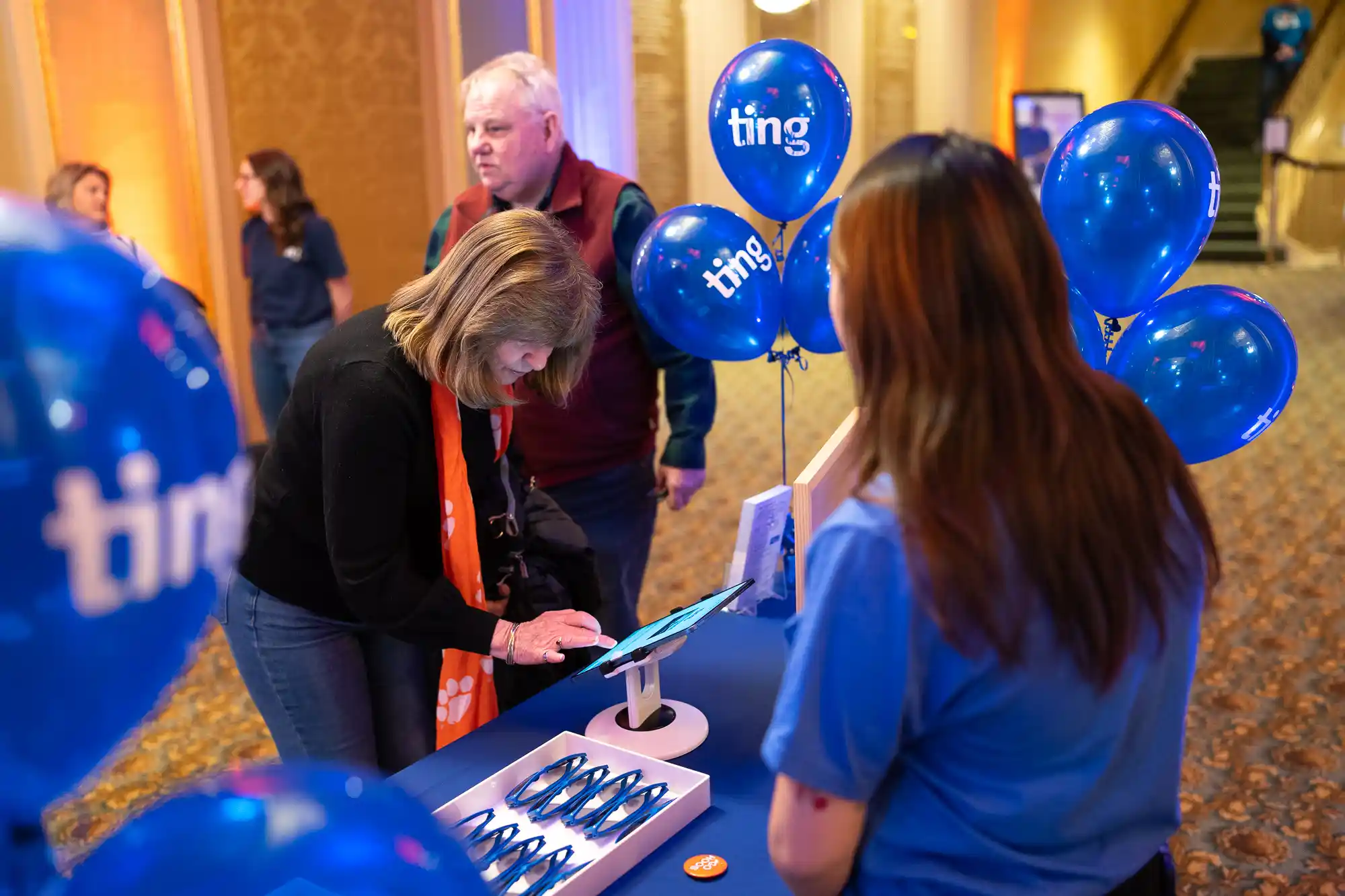 People interacting at a ting booth with blue balloons, a touch screen device, and promotional materials in an indoor setting.