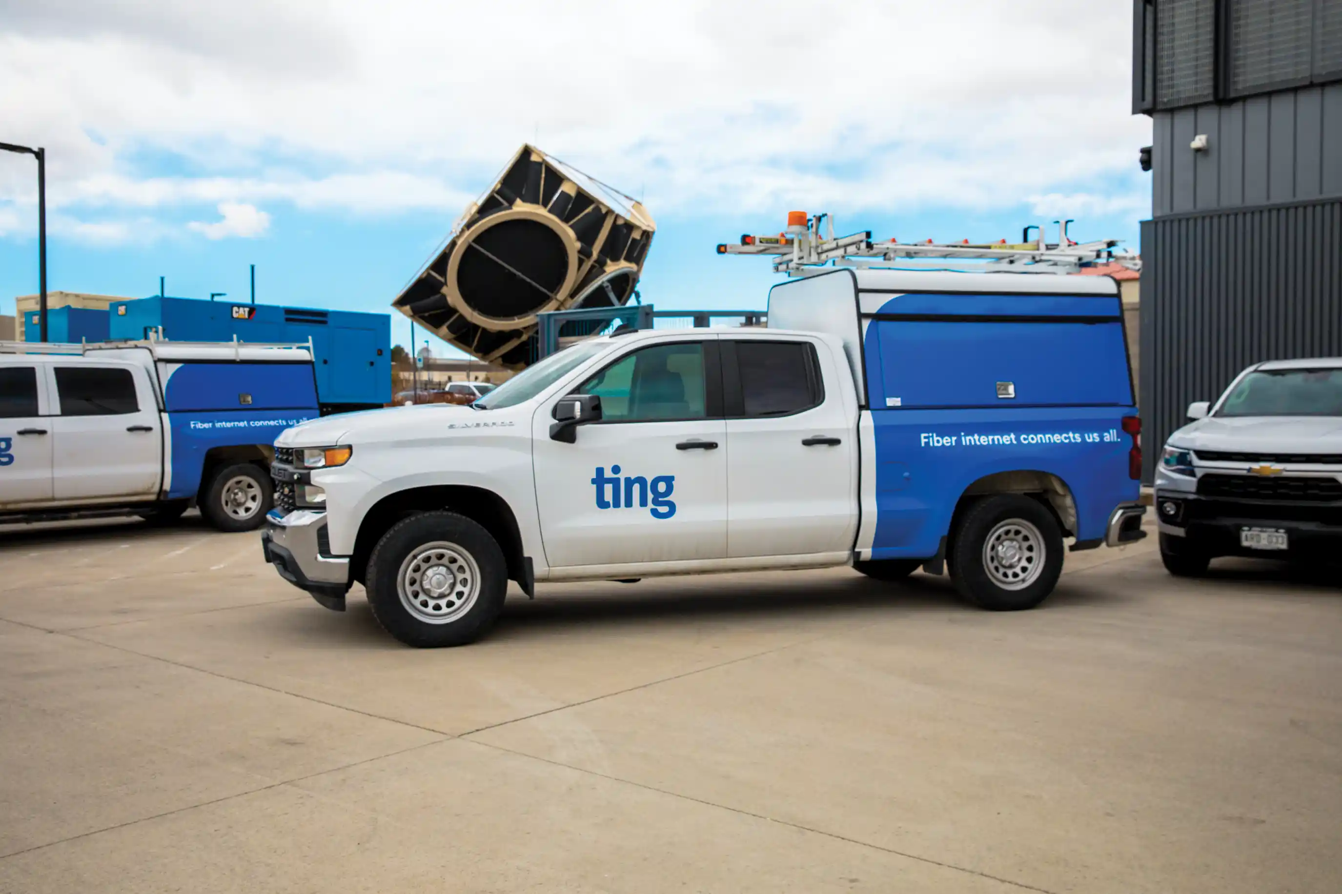 White and blue service truck with "ting" logo, parked on a concrete lot. Other vehicles and industrial equipment are visible in the background.