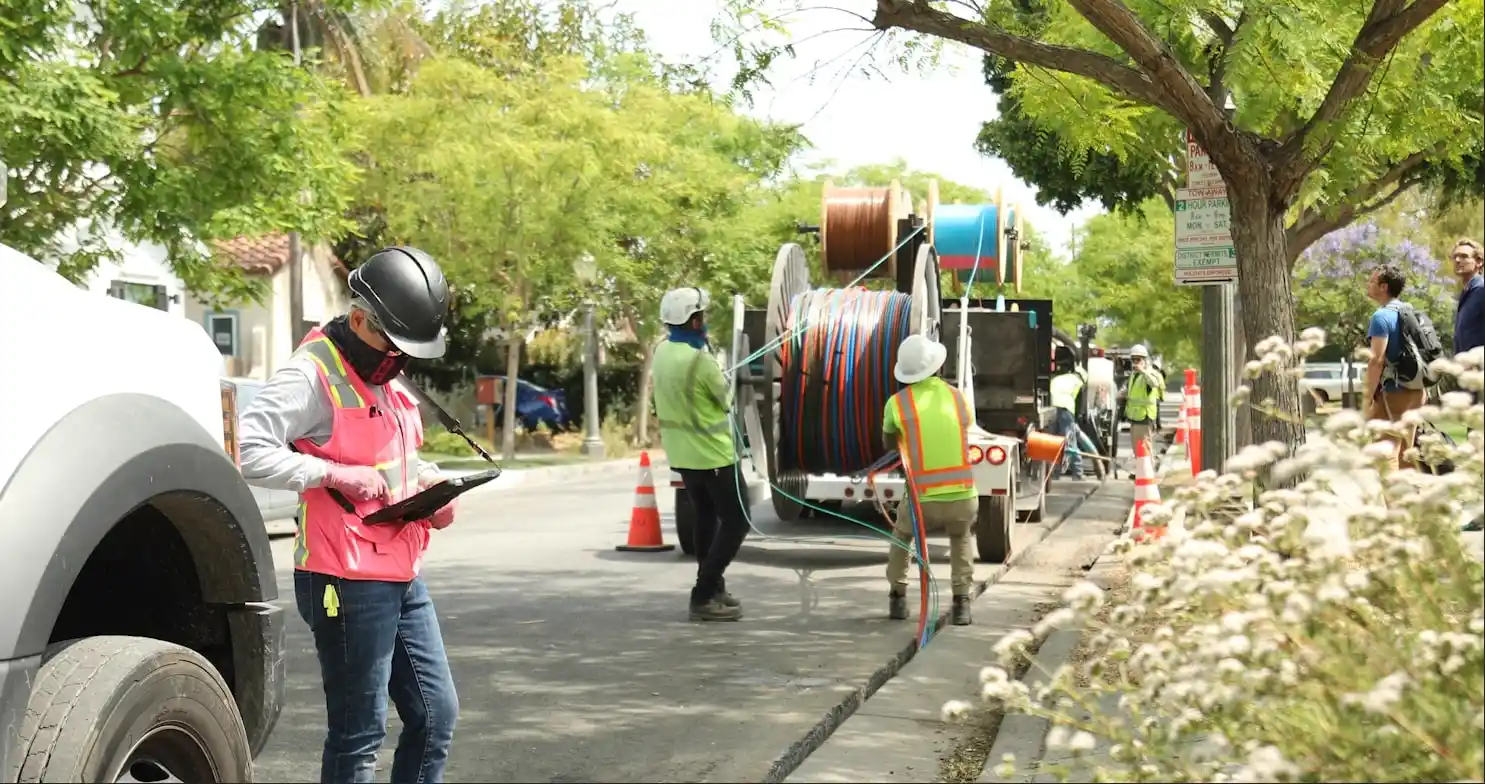 Workers wearing safety gear install cables on a residential street. Equipment and traffic cones are set up around the work area.