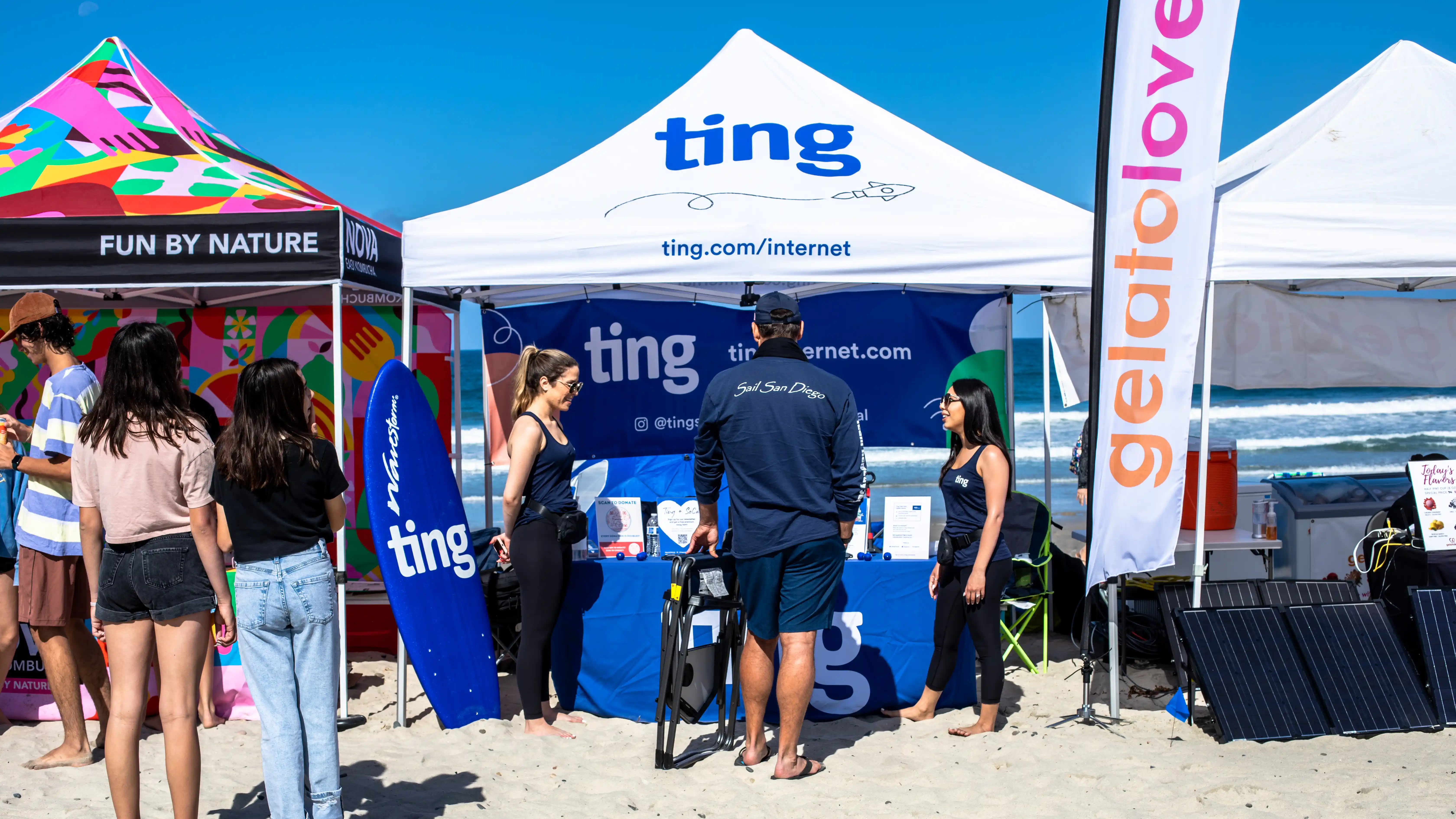 People visit a booth set up on a sunny beach with a "ting" tent and banners.