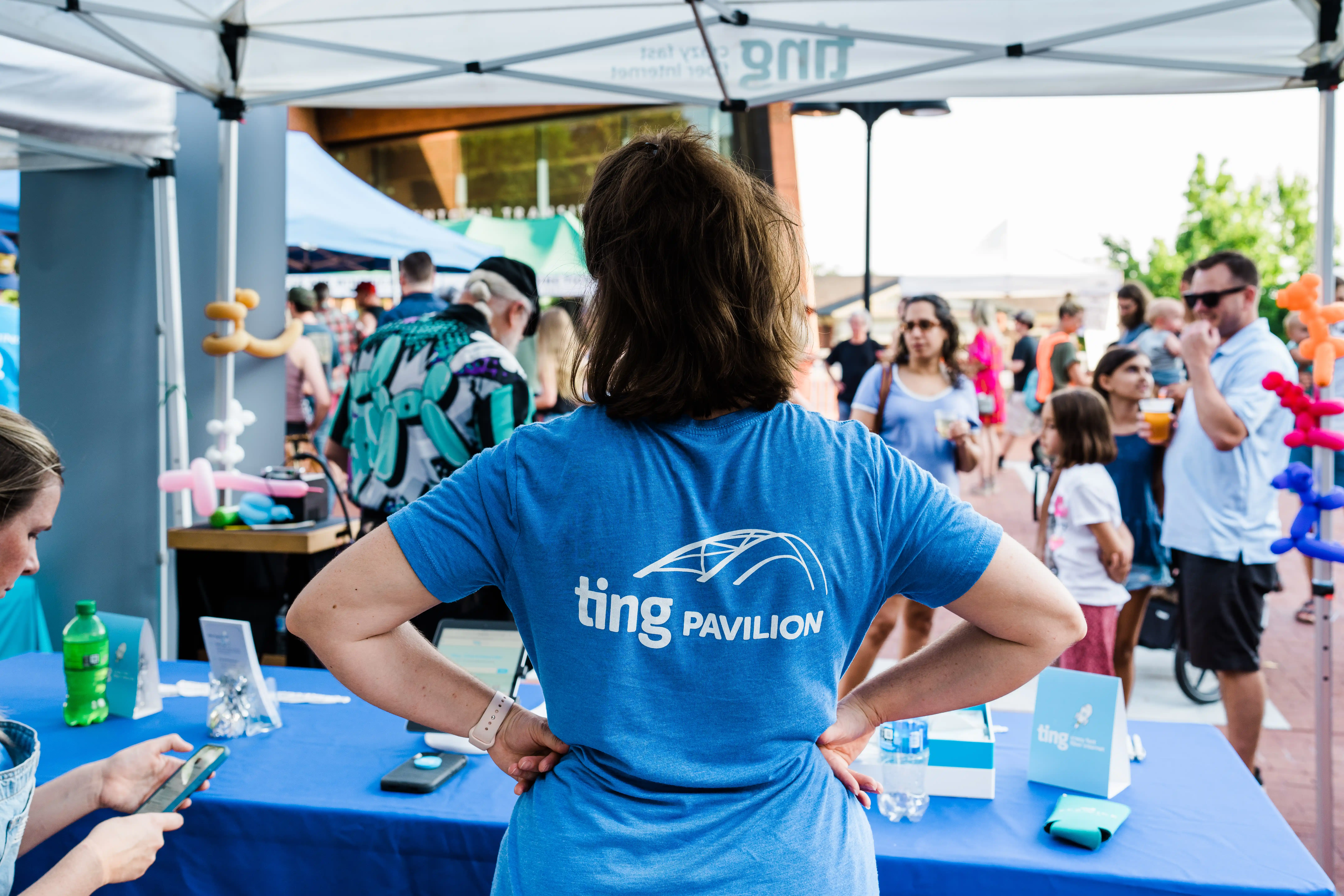 Person in a blue "Ting Pavilion" shirt standing with hands on hips at an outdoor event booth, surrounded by people, tables, and colorful balloons.
