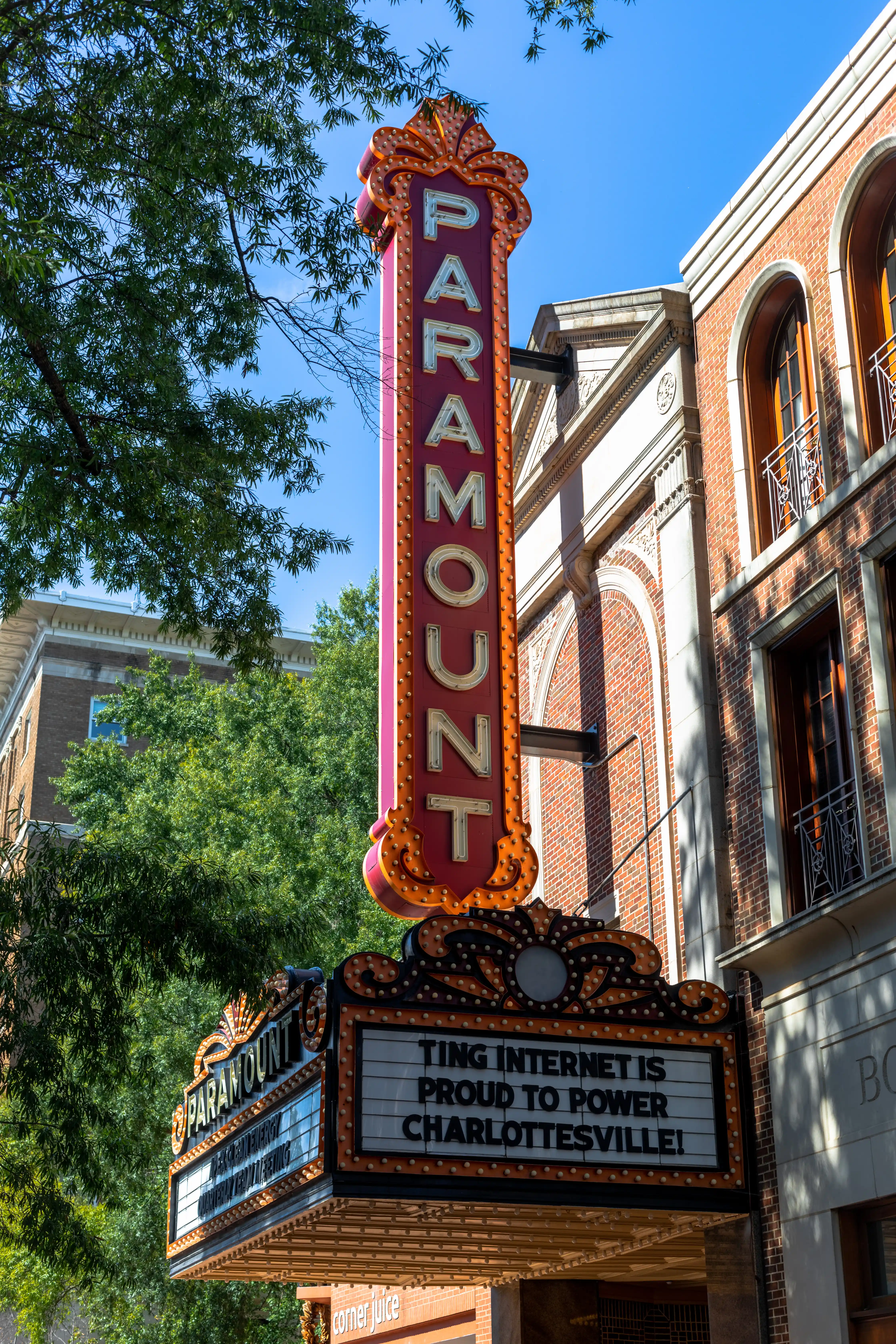 Vertical marquee sign for Paramount Theater in Charlottesville with scrolling text that reads, "Ting Internet is proud to power Charlottesville," set against trees and a brick building.