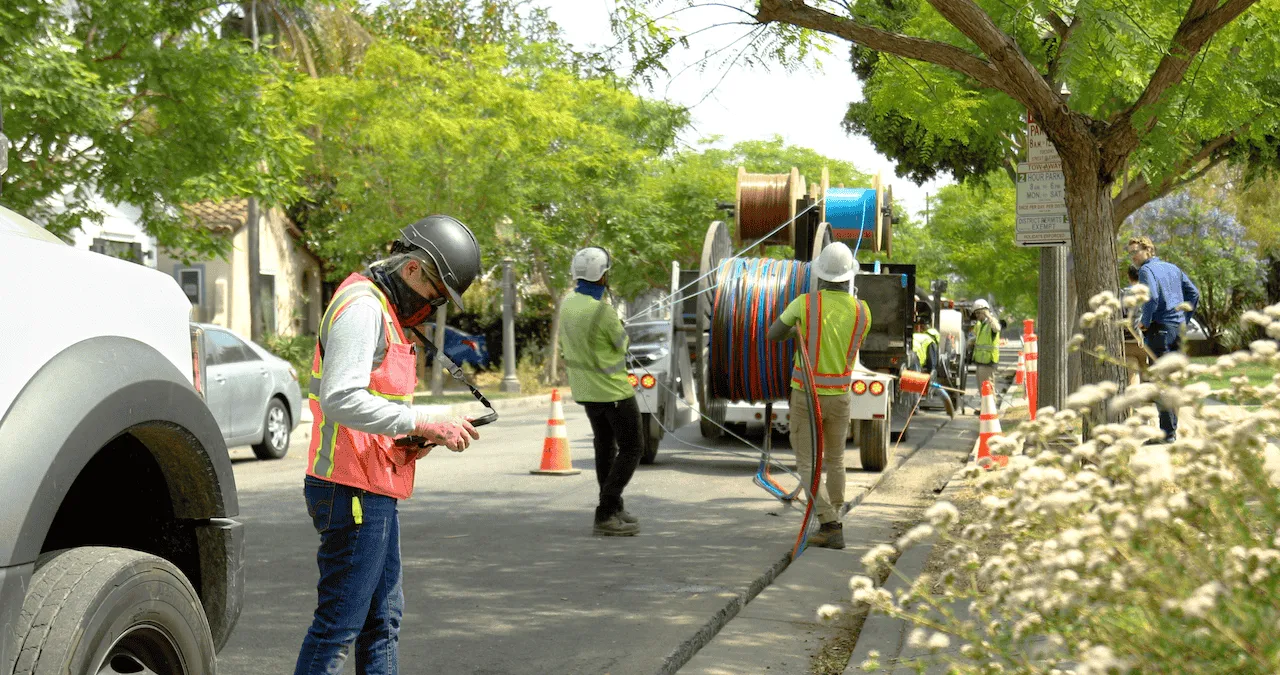 Workers wearing safety gear install cables on a residential street. Equipment and traffic cones are set up around the work area.