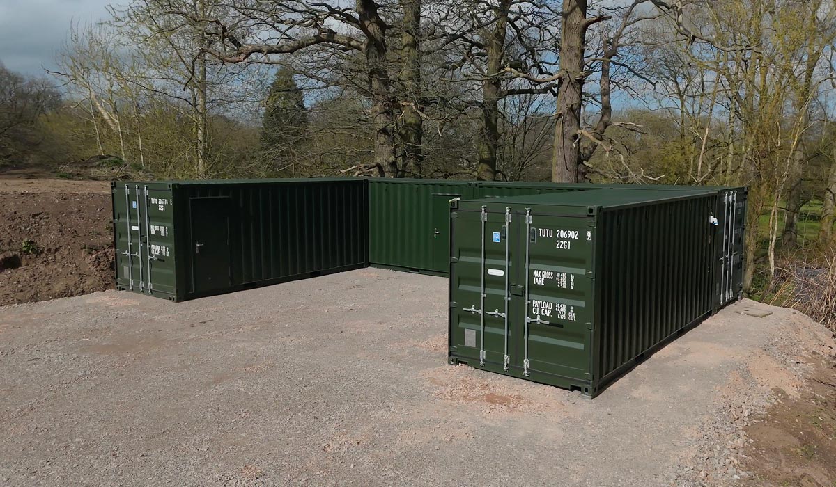 Insulated green 20ft containers with pedestrian access doors forming an open-ended storage compound beside woodland