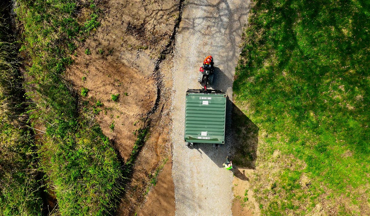 Aerial view of a green shipping container being moved along a narrow gravel track to the installation site