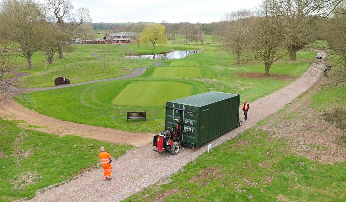 Green 20ft shipping container being delivered along a gravel path across a golf course, with the clubhouse in the background