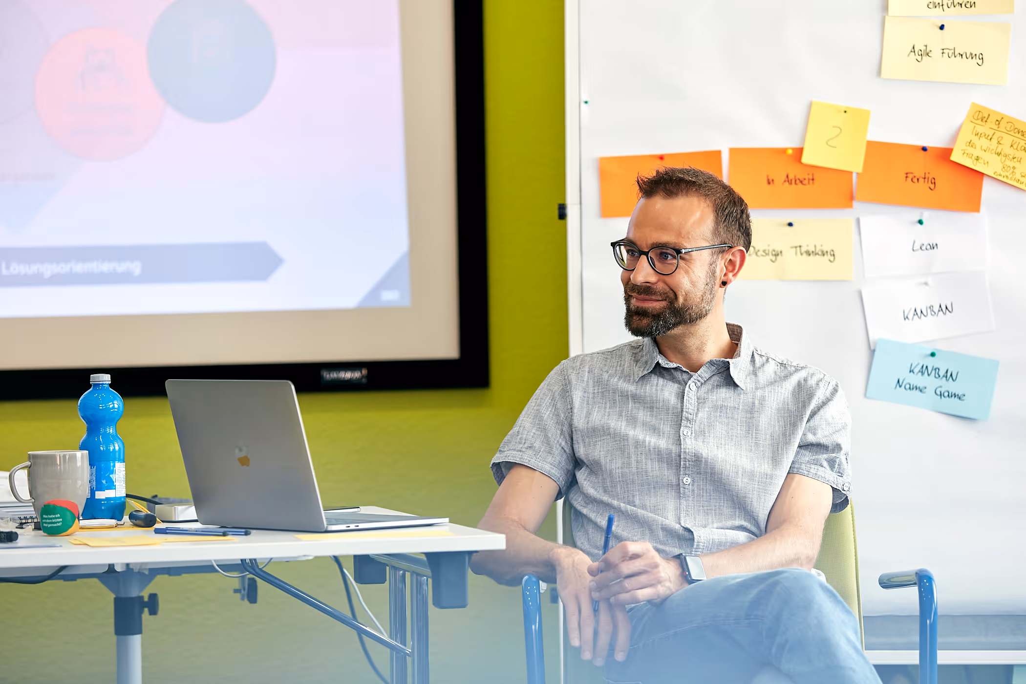 Mann mit Brille und Bart sitzt lächelnd auf einem Stuhl vor einem Whiteboard mit farbigen Haftnotizen.
