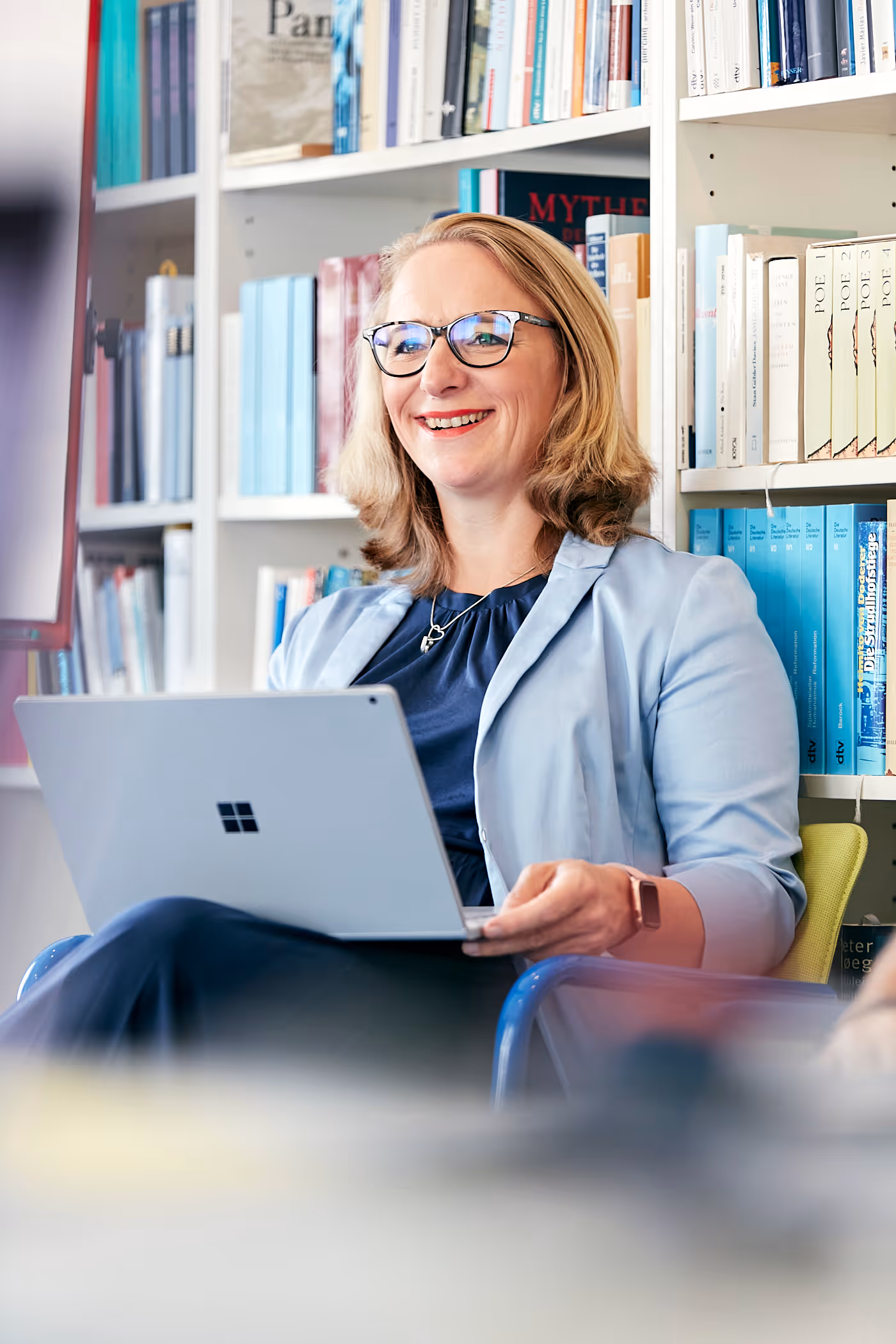 Lächelnde Frau mit Brille sitzt vor einem Bücherregal und hält ein Microsoft Surface Notebook.