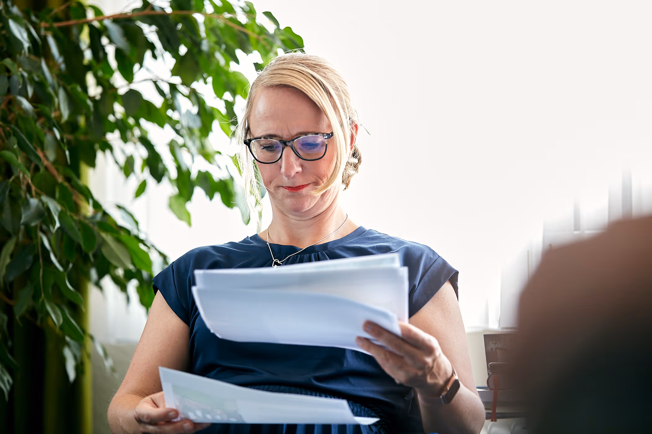 Frau mit Brille liest aufmerksam Dokumente in einem hellen Raum mit Pflanzen im Hintergrund.