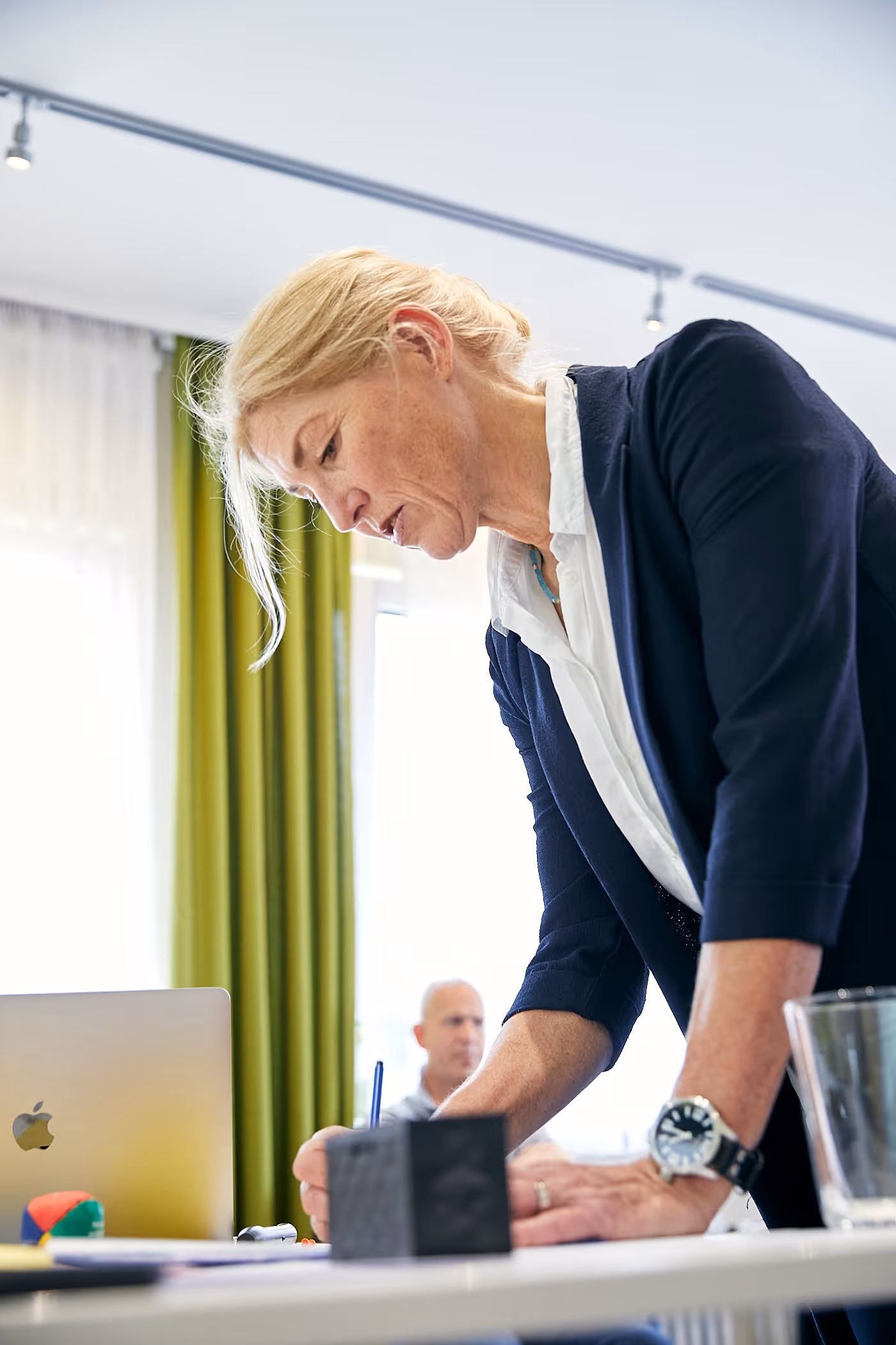 Frau beugt sich über einen Tisch und schreibt, im Hintergrund ein Mann, heller Raum mit grünen Vorhängen.