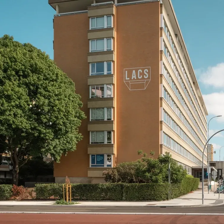 Multi-story brick building with LACS sign, surrounded by green trees and hedges on a sunny day.