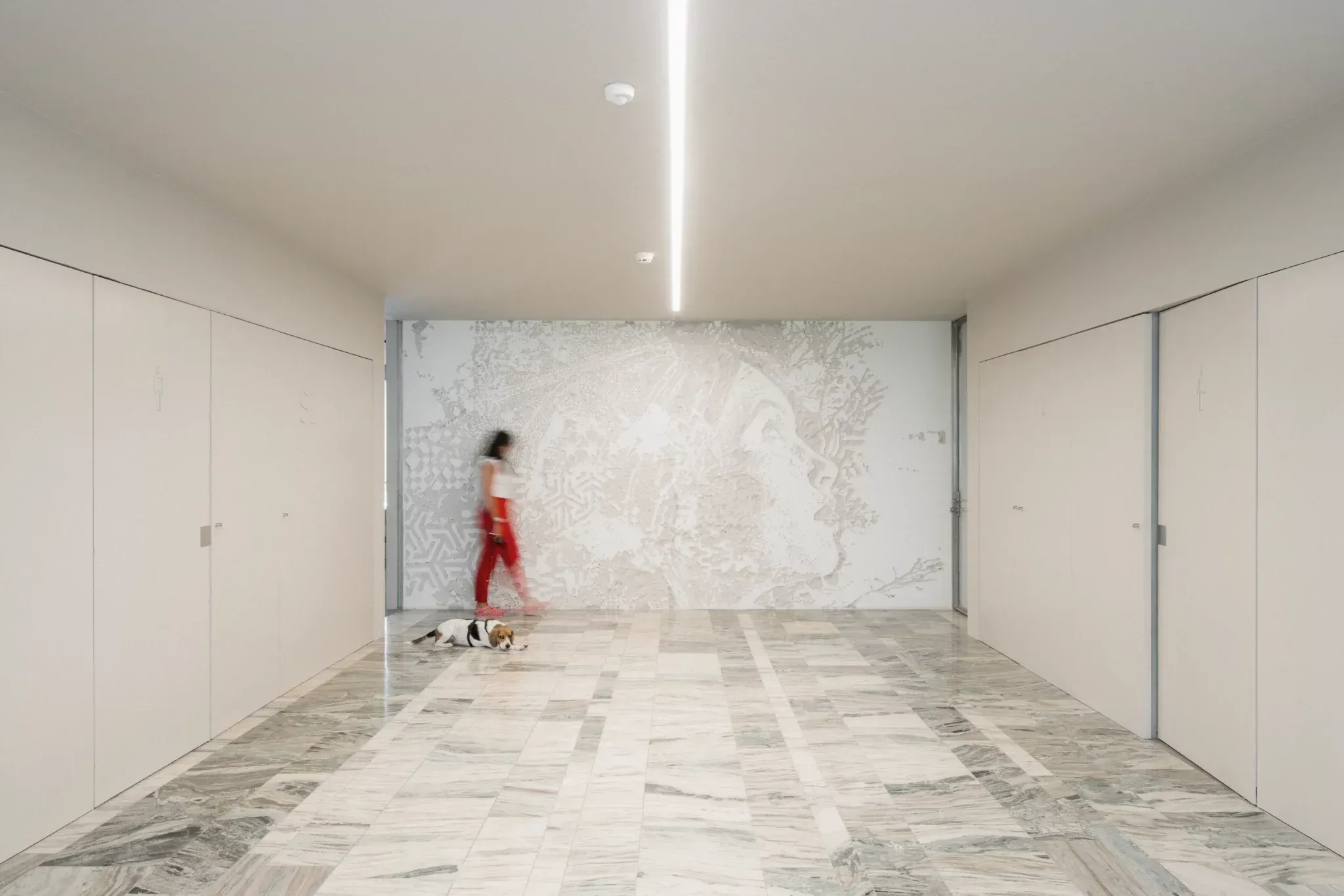 Woman walking a dog on a leash in a spacious hallway with marble floor and art on the wall.